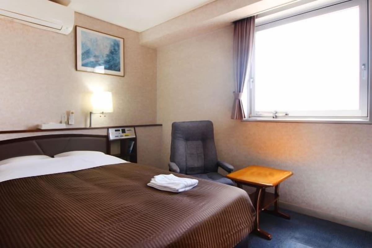 A hotel room with a large bed, a desk, a chair, a small table, a lamp, and a window with curtains.