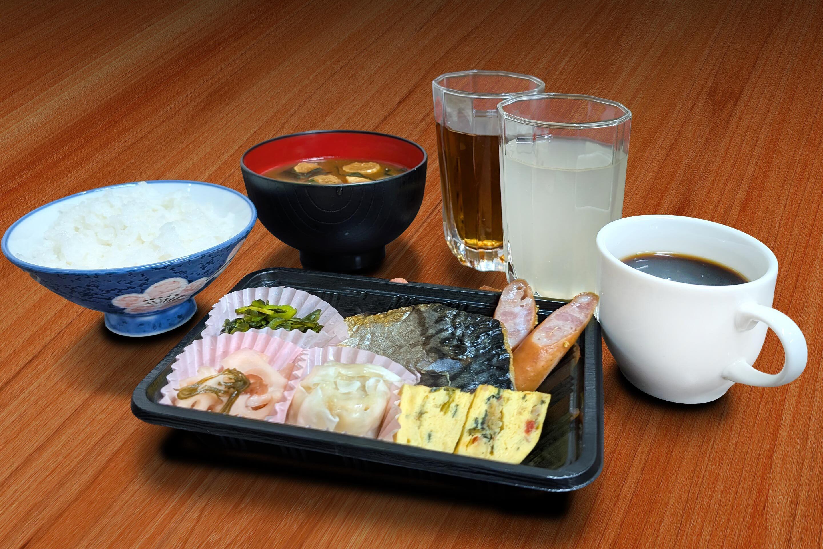 Bandeja de comida con arroz, sopa y varios platos, acompañada de dos vasos de agua y una taza de café sobre una mesa de madera.