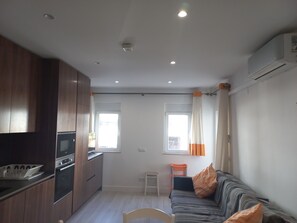Un espace de vie compact comprenant un coin cuisine, un canapé avec des coussins, une petite table et une fenêtre avec des rideaux.