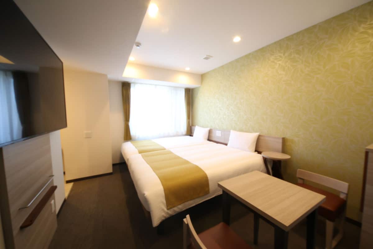 A hotel room with a large bed, a desk, and a chair. There is a window with curtains and a television on the left side.