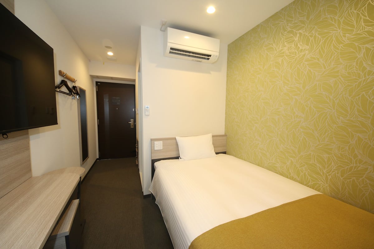 A hotel room with a bed, a desk, a TV mounted on the wall, and an air conditioning unit.