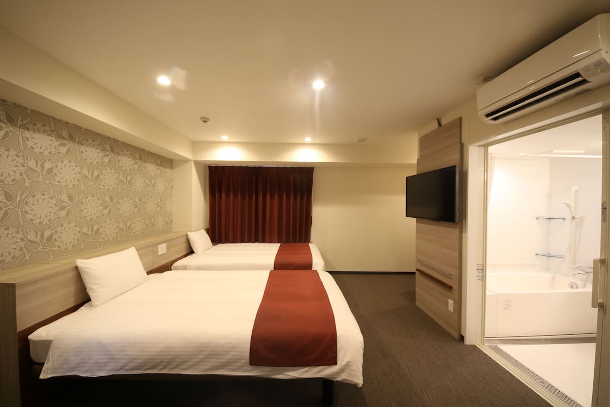 A hotel room with two beds, a TV, an air conditioning unit, and a shower area.
