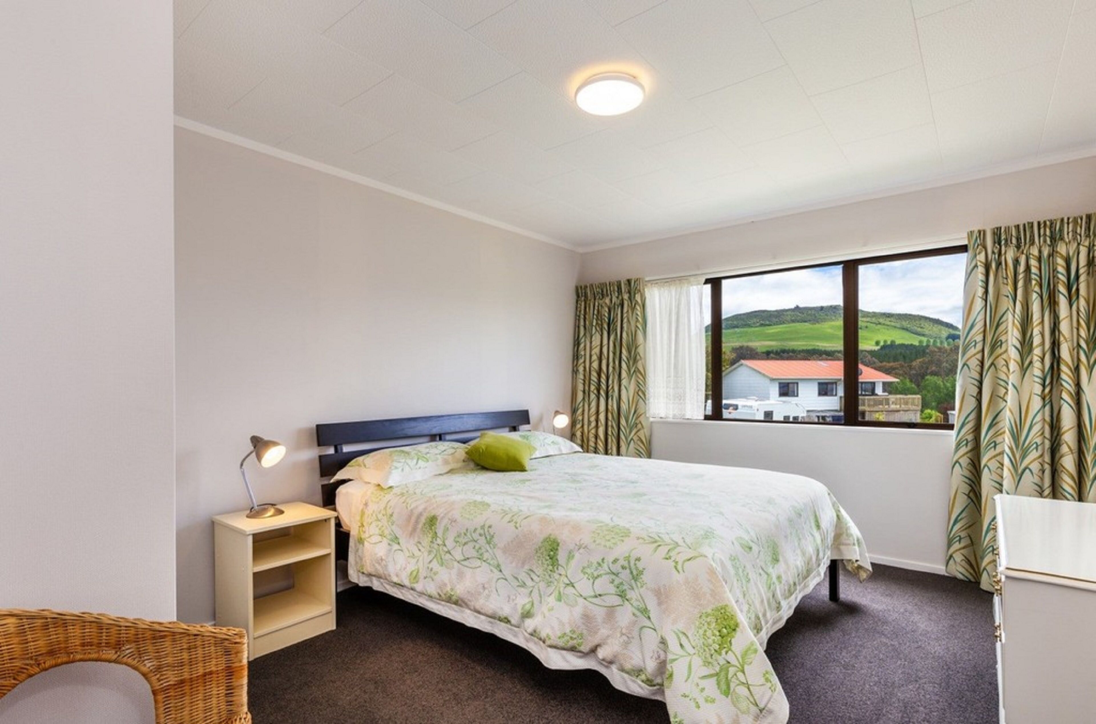 A bedroom with a bed, a bedside table, a lamp, a window with curtains, and a view of a house and landscape.