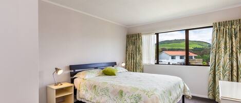A bedroom with a bed, a bedside table, a lamp, a window with curtains, and a view of a house and landscape.