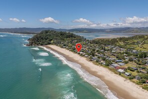 An aerial view of a coastal area with a beach, houses, and a forested hill. - Poppy's Beach House - Beachfront Whangapoua Home (Whangapoua)