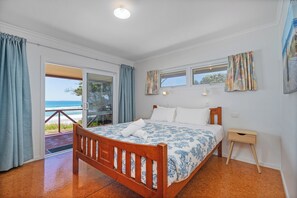 A bedroom with a wooden bed, a small bedside table, and a view of the beach through a sliding glass door. - Poppy's Beach House - Beachfront Whangapoua Home (Whangapoua)