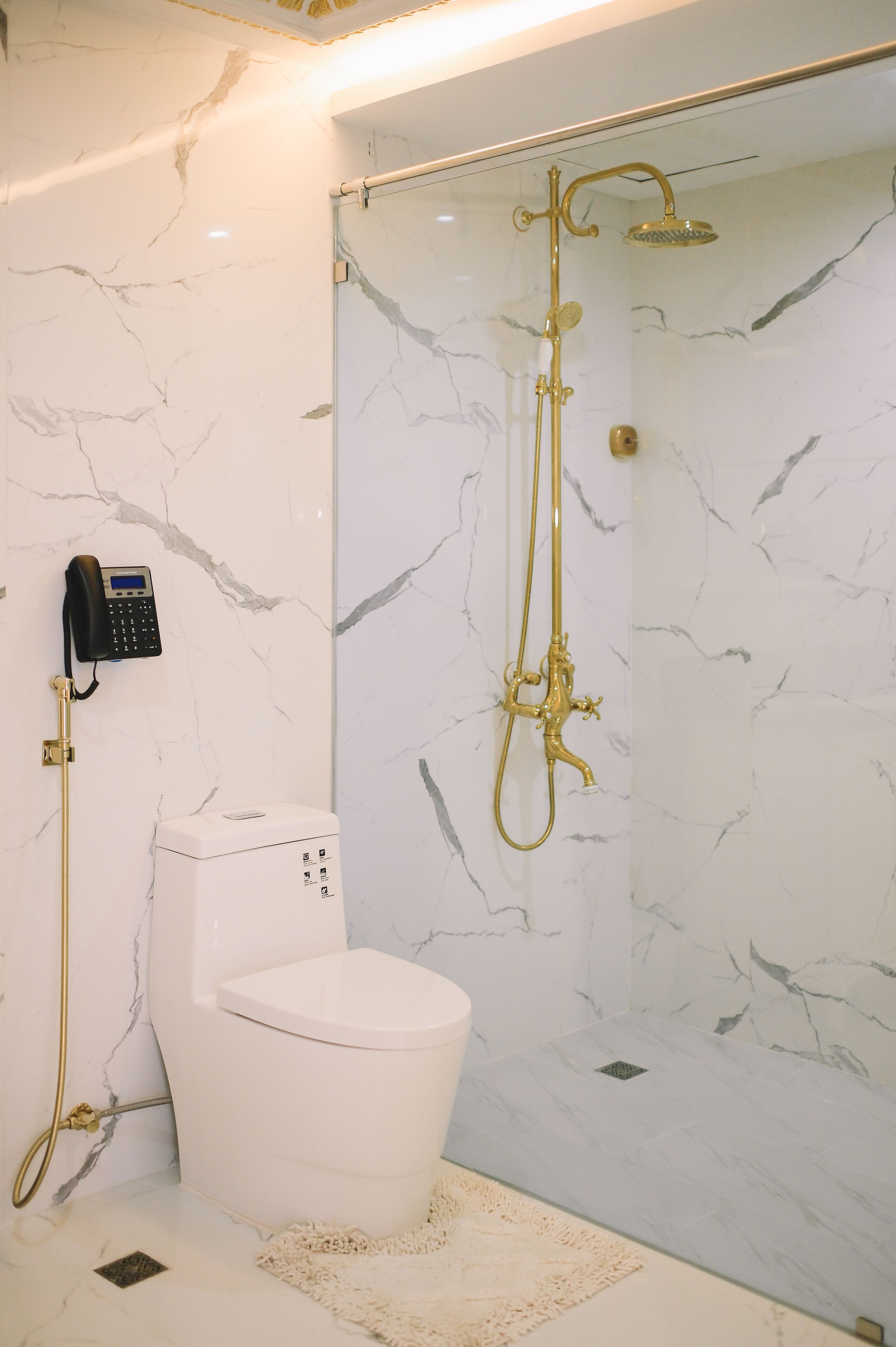 A bathroom with a marble wall, a white toilet, a gold shower system, and a telephone mounted on the wall.