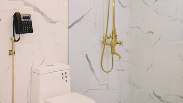 A bathroom with a marble wall, a white toilet, a gold shower system, and a telephone mounted on the wall.