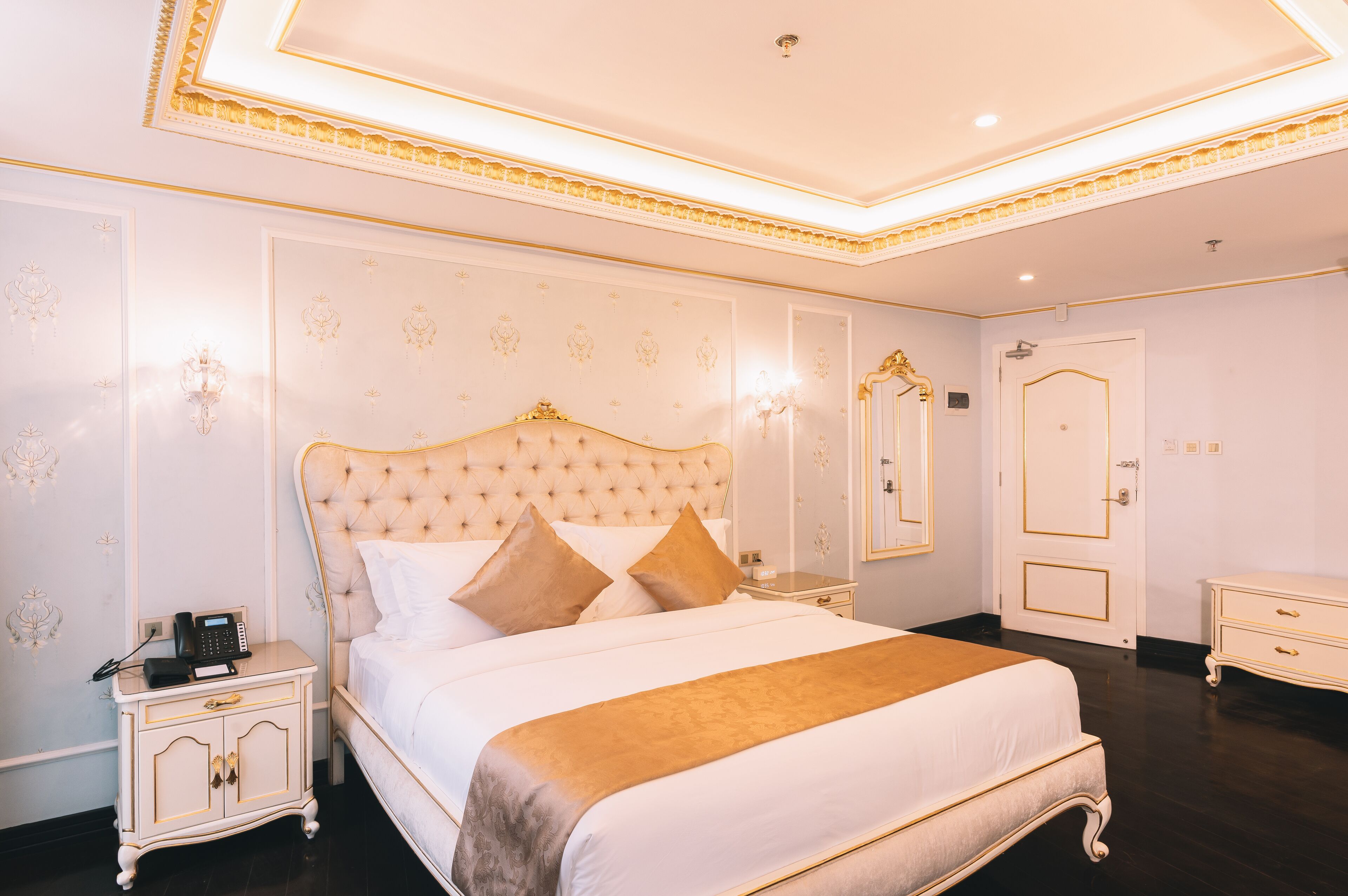 A luxurious bedroom with a tufted headboard, a large bed with white linens and a gold throw, a bedside table with a telephone, and a nightstand with a lamp.