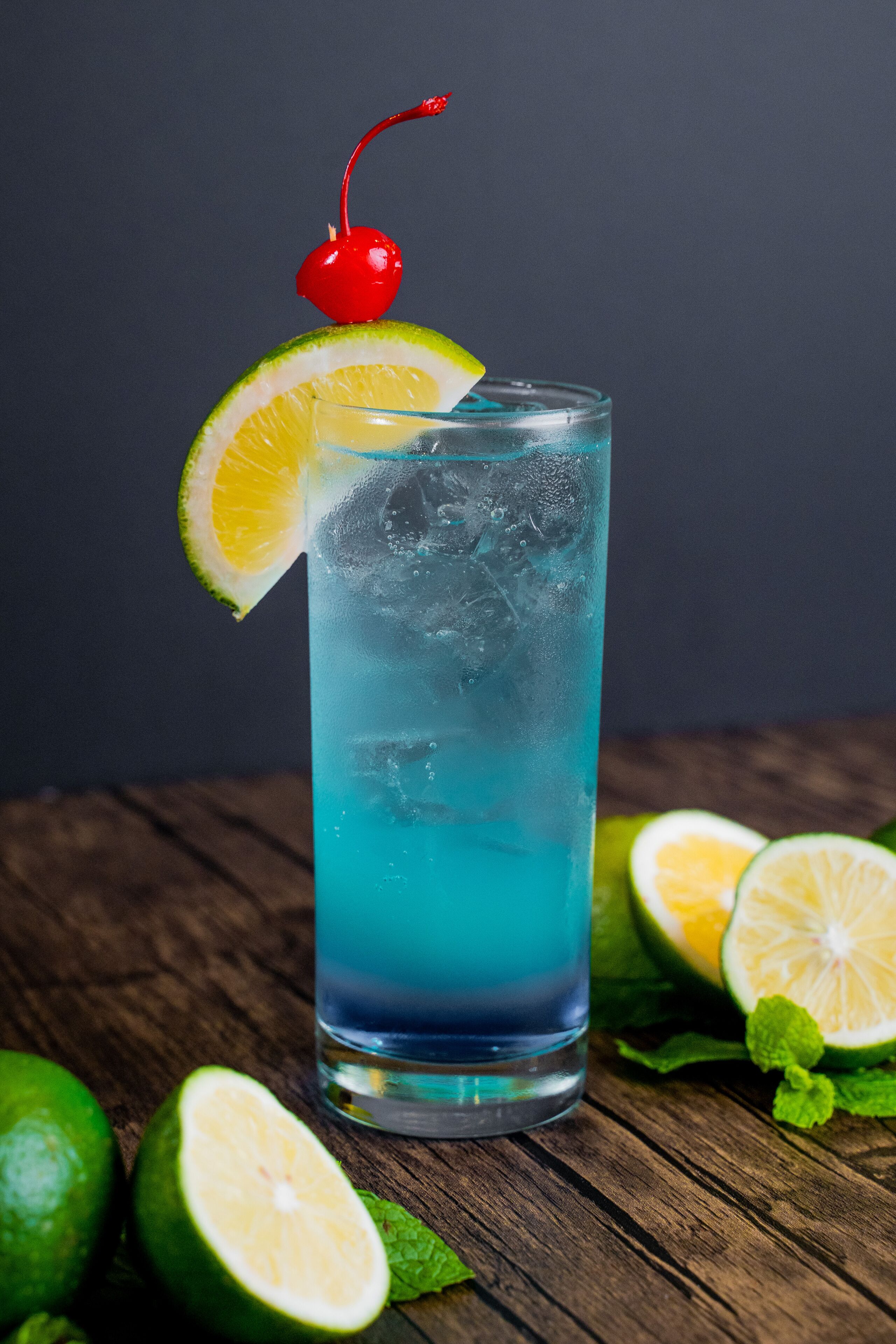 Un grand verre contenant une boisson bleue, décoré d’une cerise et d’une tranche de citron vert, entouré de citrons verts entiers et coupés en deux sur une surface en bois.