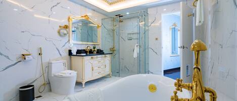 Un baño lujoso con piso de mármol, una bañera blanca, un cabezal de ducha dorado y un tocador con espejo dorado.