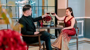 Un hombre y una mujer sentados a una mesa con una botella y copas, en una habitación con una decoración moderna y un ventanal grande.