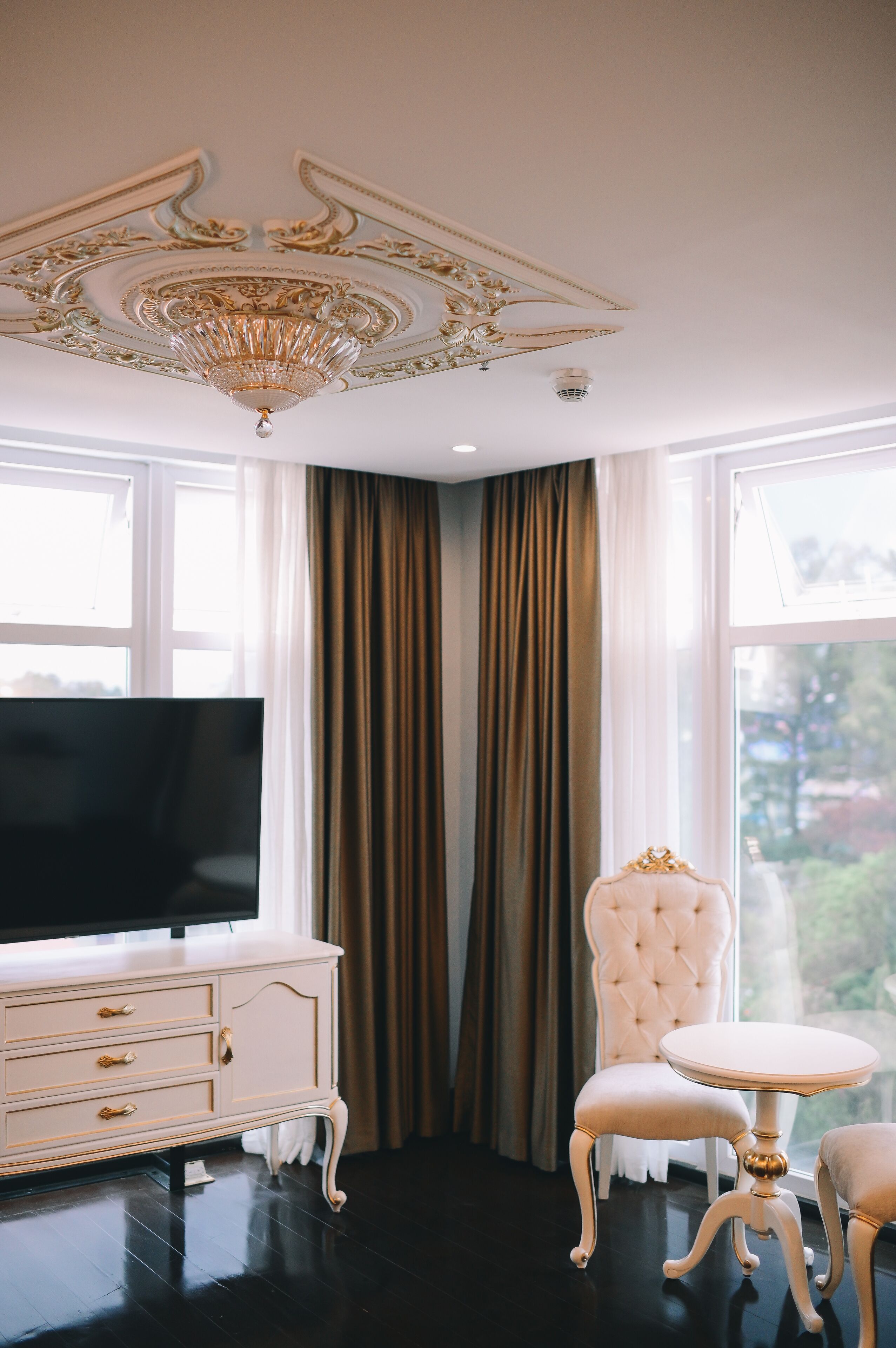 A room with a large flat-screen TV, a white ornate cabinet, a tufted chair, and a small round table.