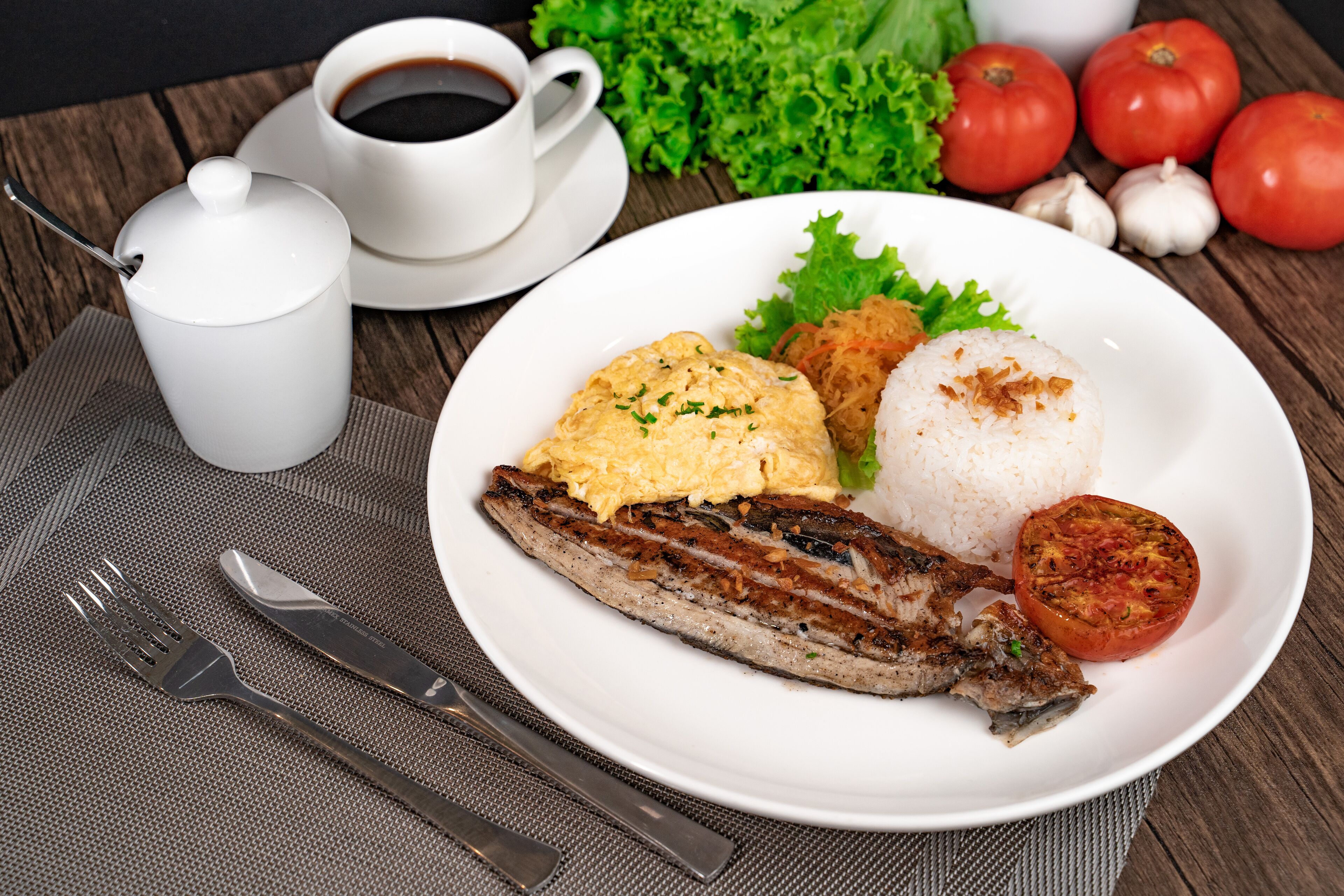 A plate with rice, scrambled eggs, a grilled fish, and a grilled tomato on a wooden table.