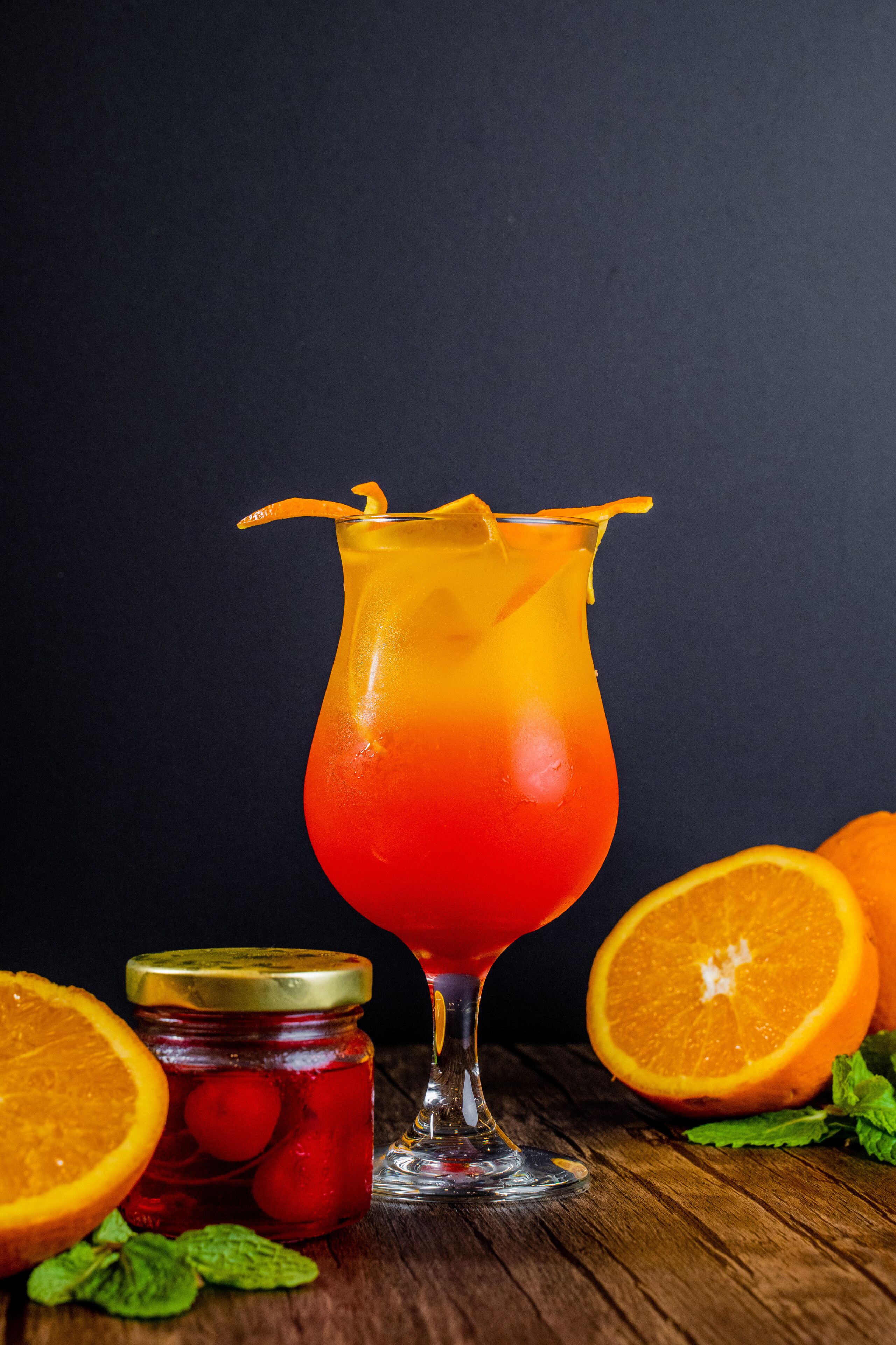 Un vaso de cóctel con un degradado de líquido rojo a naranja, una rodaja de naranja y un tarro de fruta en conserva sobre una superficie de madera.