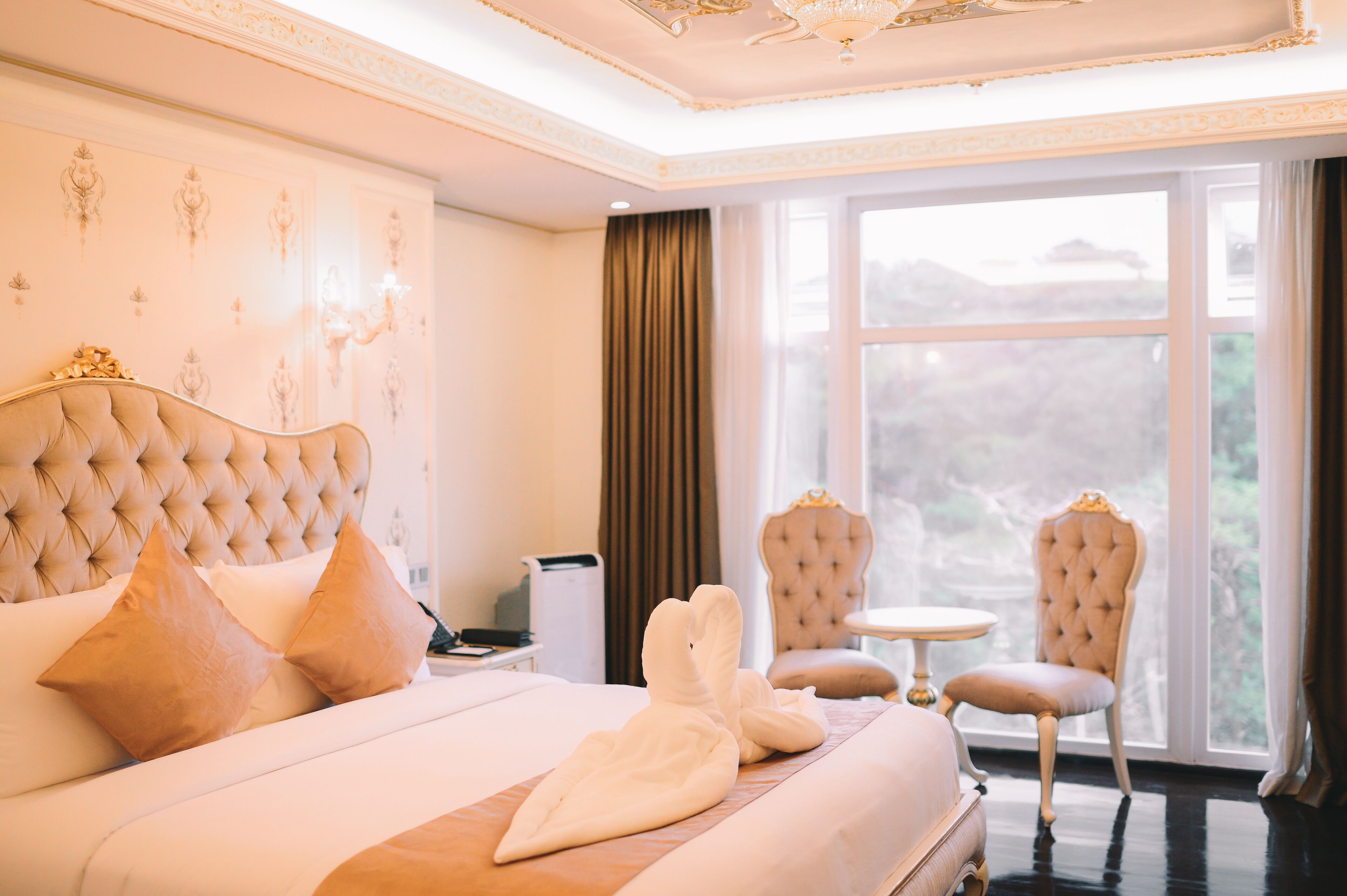 A luxurious hotel room with a large bed, two armchairs, a small round table, and a view of the outdoors through a large window.