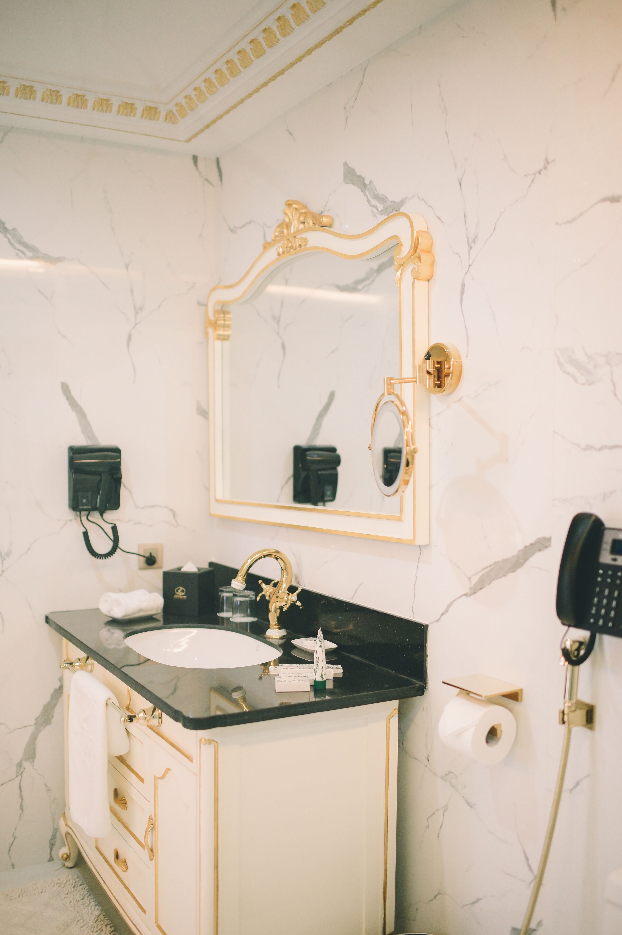 Ein Badezimmer mit Marmorwand, einem goldfarbenen Spiegelrahmen, einer schwarzen Arbeitsplatte, einem goldenen Wasserhahn und einem schwarzen Telefon.