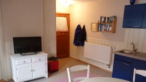 Una habitación pequeña y bien iluminada con una mesa y sillas blancas, un televisor sobre un mueble blanco, un radiador, un lavabo y un armario azul lleno de libros.