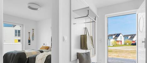 A modern bedroom with a bed, a hanging bag, and a view of a residential area through an open door.