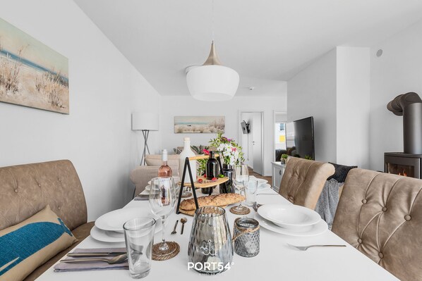 Un comedor con una mesa blanca dispuesta para cuatro personas, con una lámpara colgante moderna, un ramo de flores y una variedad de vajilla.