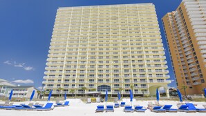 A large hotel with multiple floors, a central entrance, and a beach area with sun loungers and umbrellas. - Renovated 1BR Celadon Condo | Gulf Views, Balcony, Bunks + Beach Chairs Included! (Panama City Beach)