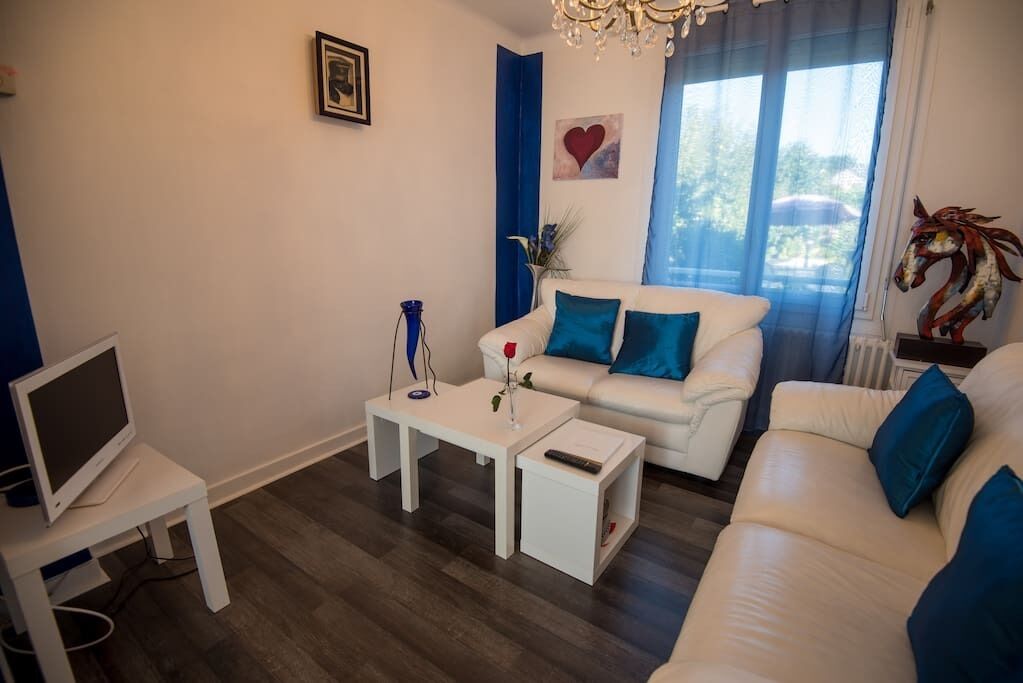 A modern living room with a white sofa, a coffee table, a TV on a stand, and a window with blue curtains.