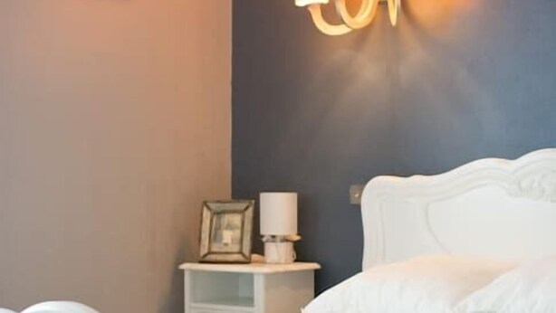 A bedroom with a white bed, a nightstand with a lamp, and a wall-mounted light fixture. - entre parc et Haras - Hennebont (Hennebont)