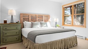 Una camera da letto con una testiera in legno, un letto con biancheria bianca, un comò verde, due lampade sui comodini e una finestra vista alberi.