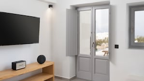 A modern living room with a flat-screen TV, a wooden entertainment unit, a small round table with a bowl of fruit, a grey door, and a window.