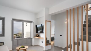 A modern living room with a large window, a flat-screen TV, a wooden table with fruit, and a staircase with wooden railings.