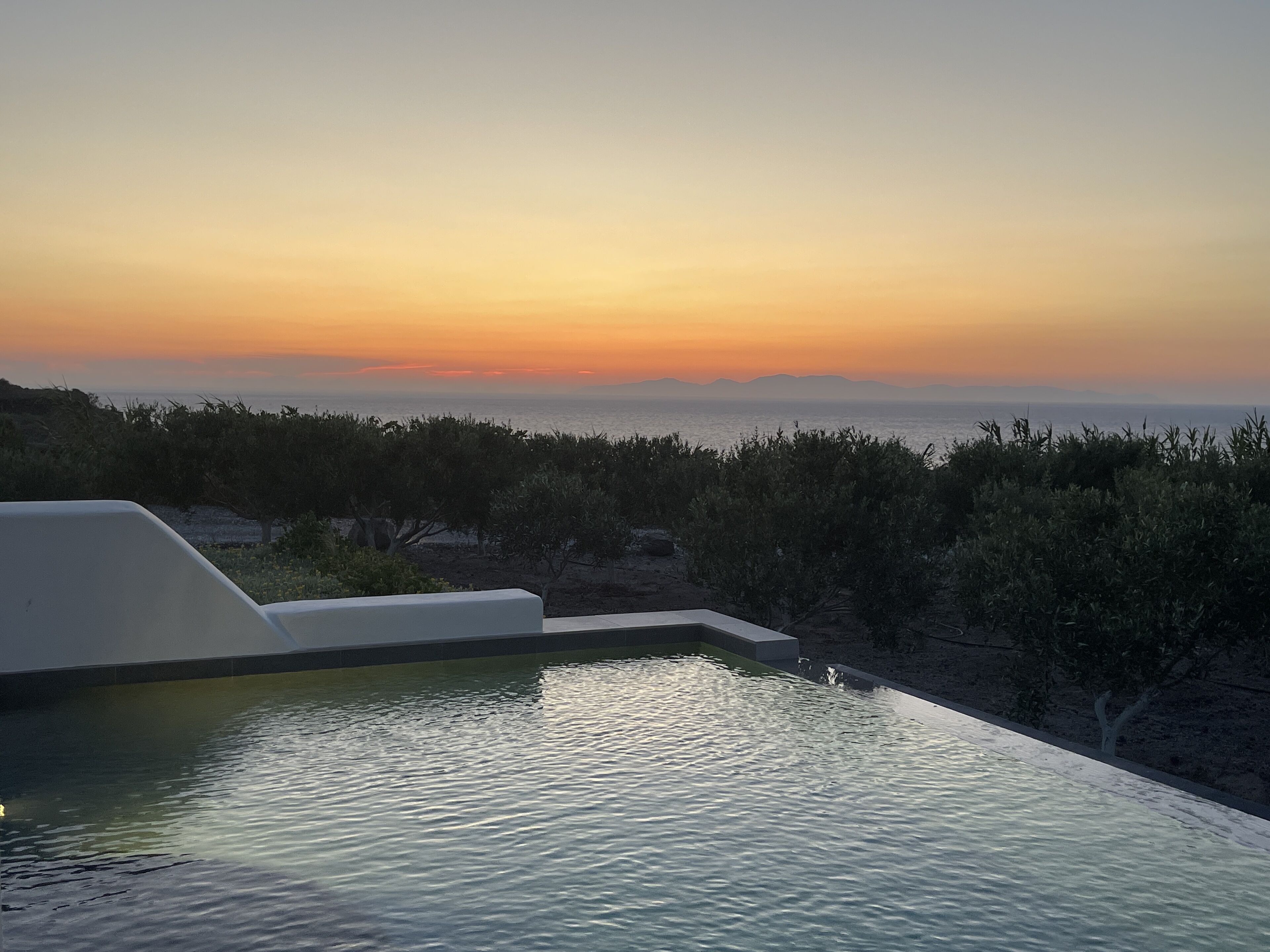 Une piscine offrant une vue sur le coucher de soleil sur la mer, entourée de végétation.