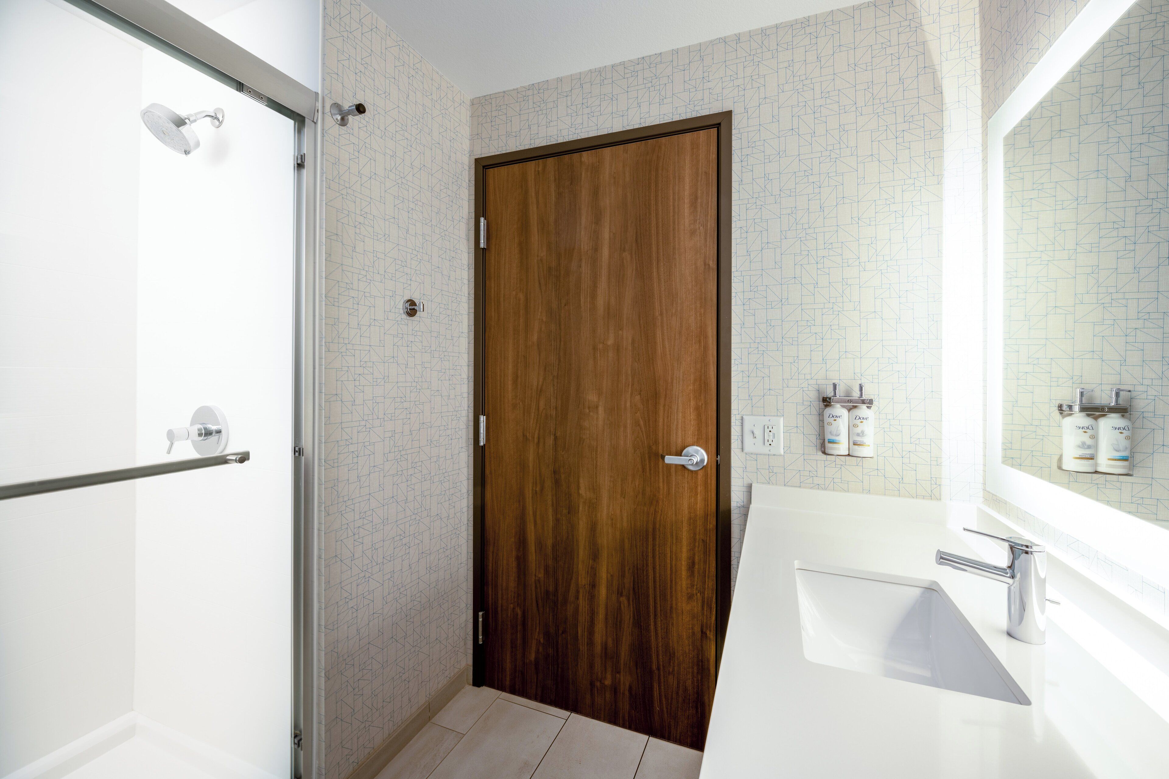 Un baño con puerta de madera, ducha con puerta de cristal y lavabo con espejo encima.