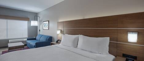 A modern hotel room with a large bed, a blue armchair, a bedside table with a lamp, and a nightstand with a clock.