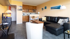 A modern living room with a black leather sofa, a white armchair, and a glass coffee table. A kitchen with wooden cabinets and a dining area with a table and chairs is visible in the background. - T2 Apartment Biscarrosse - AC, Parking, 4 guests, City Center (Biscarrosse Plage)