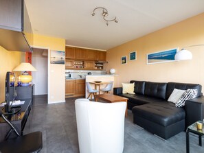 Uma sala de estar moderna com um sofá de couro preto, uma poltrona branca e uma mesa de centro de vidro. Uma cozinha com armários de madeira e um espaço de jantar com mesa e cadeiras são visíveis ao fundo.