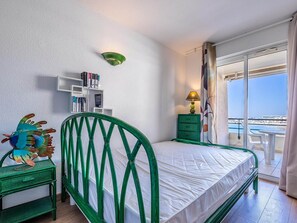Ein Zimmer mit einem Bett, einem grünen Metall-Kopfstück, einem kleinen Nachttisch, einem grünen Beistelltisch, einer an der Wand befestigten Regal mit Büchern, einer Lampe und Meerblick durch eine Glastür.
