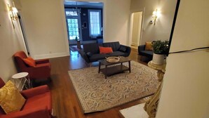 A spacious living room with hardwood flooring, a large rug, and a sofa set. There is a coffee table in the center, and a potted plant on the right.