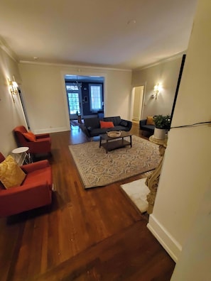 Un salón amplio con suelo de madera, un tapete grande y un conjunto de sofás. Hay una mesa de centro en el medio, y una planta en maceta a la derecha.