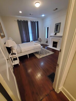 A bedroom with a bed, bedside table, lamp, and a fan. There is a fireplace and a mirror on the wall.