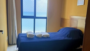 A single bed with a blue bedspread and two folded towels, a window with a mesh screen, and a wooden headboard. - Dune& Ocean Views Paradise 2 Beds in Natal (Natal)