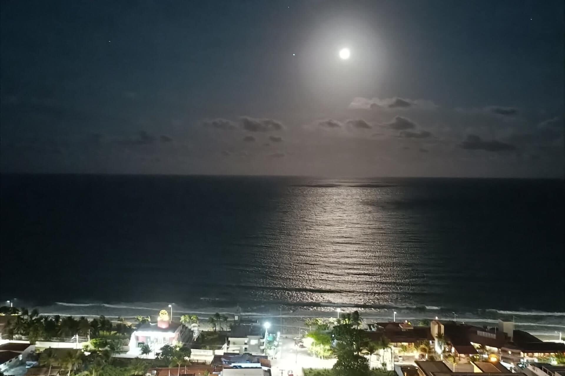 Un pueblo costero de noche con luna visible, mar en calma y edificios a lo largo de la costa.