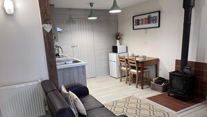 A modern living room with a black leather sofa, a dining table with chairs, a kitchen area with a sink and appliances, and a wood-burning stove. - Poachers Hut 1-bed Cottage, Sleeps 2, Pet Friendly (Newport)