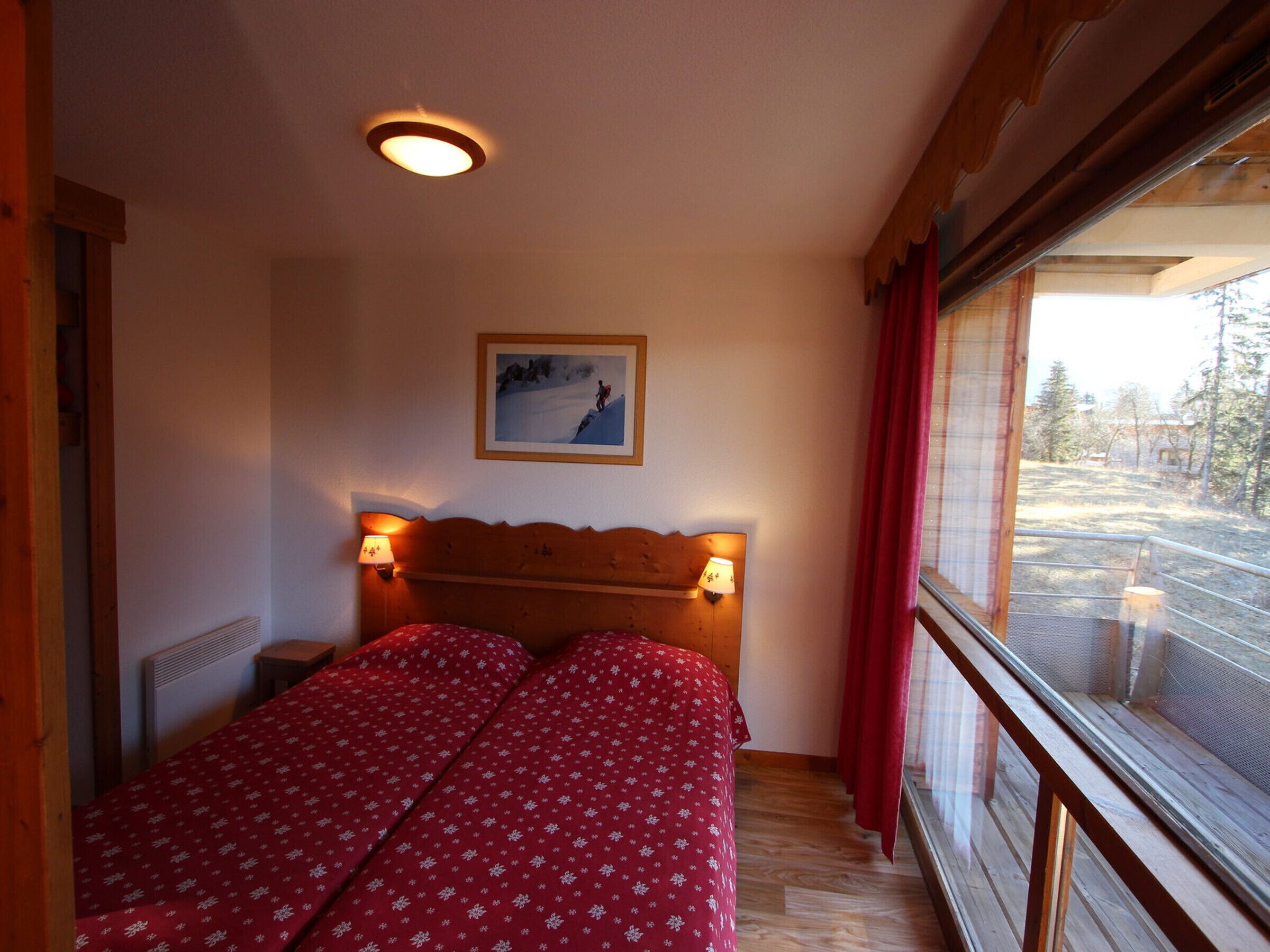 A bedroom with a wooden headboard, two beds with red floral bedding, a wall-mounted picture, and a window with a view of trees and a building.