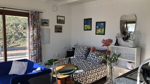 Ein modernes Wohnzimmer mit einem blauen Sofa, gemusterten Kissen, einem Esstisch mit einem grünen Handtuch und einer Küchenzeile inklusive Mixer und Kaffeemaschine.