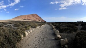 Wüstenlandschaft mit einem markanten Hügel, einem Feldweg, vereinzelter Vegetation und einem Schiffen am Horizont.
