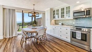 A modern kitchen with white cabinets, a stainless steel oven, and a dining area with a wooden table and wicker chairs. - Renovated Oceanfront Condo with Ground-Level Patio on Amelia Island (Fernandina Beach)