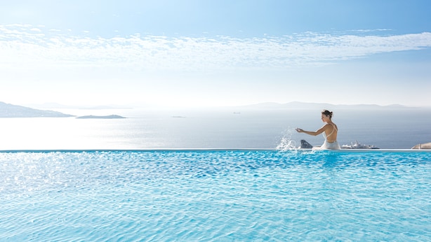 Eine Frau in einem weißen Badeanzug an einem klaren, blauen Pool mit Blick auf das Meer und entfernte Inseln.