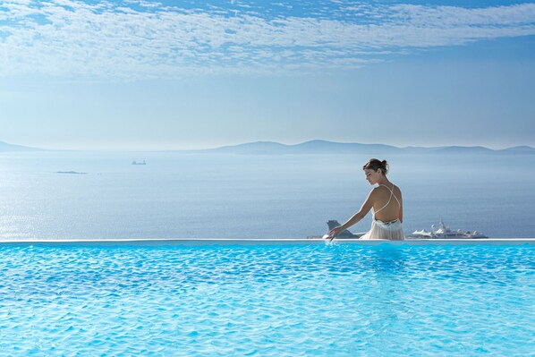 A person in a white swimsuit standing at the edge of a clear blue pool with a view of the sea and distant mountains.