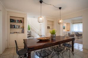 Uma área de refeições com mesa de madeira, cadeiras de arame e luminárias pendentes. Estantes à esquerda e uma escada ao fundo.