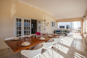 Uma sala de jantar com mesa de madeira posta para seis pessoas, cadeiras brancas e uma varanda com sofá e objetos decorativos.